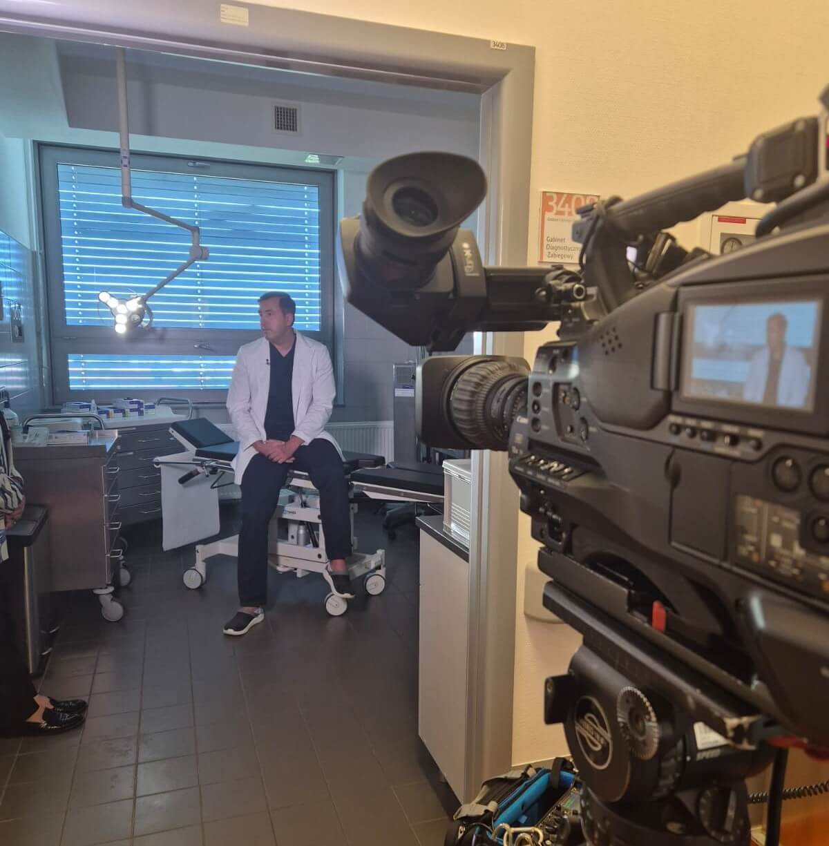 Chirurg plastyczny dr Szymon Kołacz rozmawia z dziennikarką podczas wywiadu telewizyjnego. Mężczyzna w fartuchu chirurgicznym siedzi na stole operacyjnym, uśmiecha się i gestykuluje. W tle widoczne są profesjonalne kamery oraz oświetlenie.